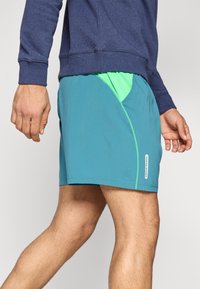 Τυρκουάζ σορτς με πράσινο διακριτικό από δίχτυ· ελαφρύ ύφασμα, κανονική εφαρμογή και λογότυπο στη ραφή στο πλάι. Ιδανικά για αθλητική χρήση.