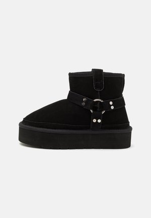 Botte cheville en daim noir avec semelle plateforme épaisse, sangle décorative, anneau en métal et détails en rivets argentés.