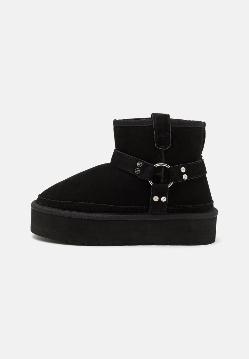 Botte cheville en daim noir avec semelle plateforme épaisse, sangle décorative, anneau en métal et détails en rivets argentés.