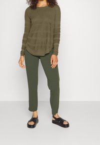 Frau trägt ein olivgrünes, geripptes Langarmshirt, dunkelgrüne Hose und schwarze Plateausandalen, steht vor einem einfarbigen Hintergrund.