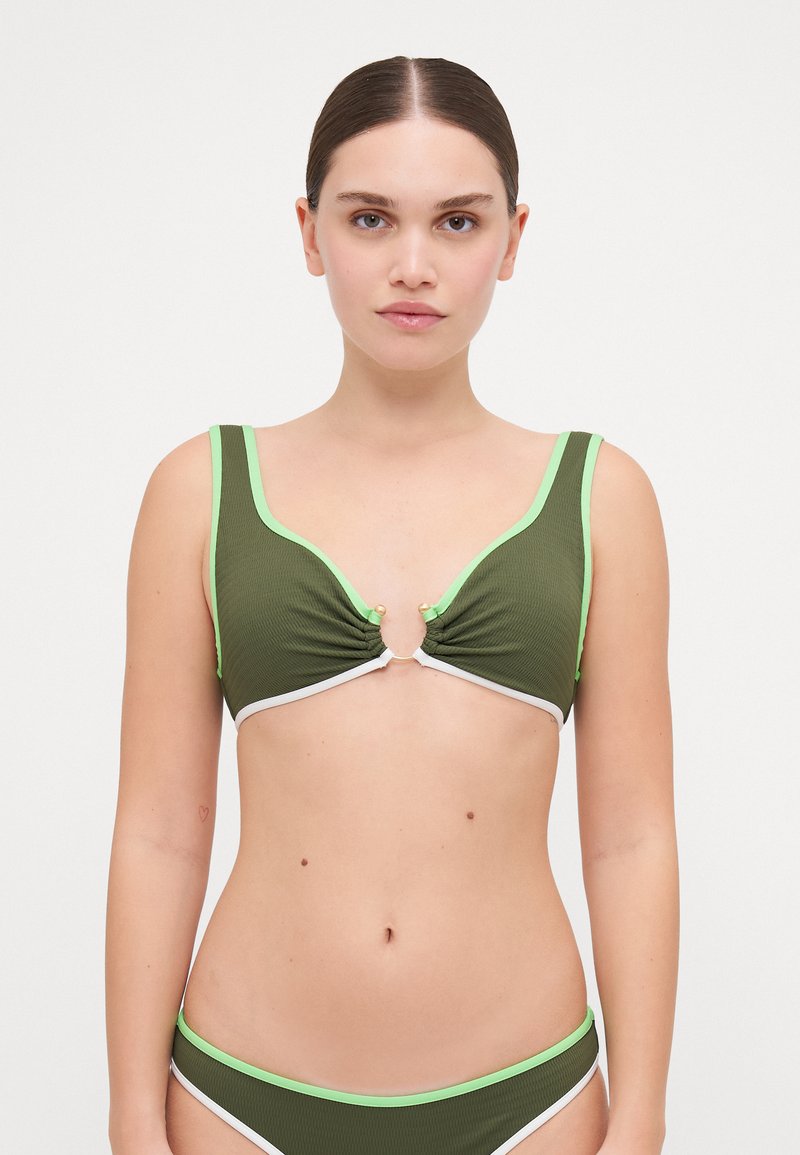 Donna che indossa un bikini verde oliva con rifiniture verde chiaro e bianche, in piedi contro uno sfondo bianco semplice, rivolta in avanti.