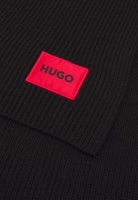 Beanie en tricot noir avec texture côtelée. Présente un patch rectangulaire rouge avec "HUGO" en lettres noires en gras. Fini en tissu doux.