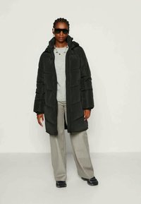 Melnā pūkainā virsjakā ar garām piedurknēm, kapuci un quiltoto rakstu. Valkāta virs pelēkā krekla, kopā ar svītrainām bēšām biksēm un tumšām kurpēm.