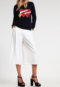 Marineblauer Pullover mit rotem und goldenem Herz-Graphic und dem Text "LOVE TOMMY". Weiße weit geschnittene Hosen und schwarze Blockabsatzsandalen runden den Look ab.