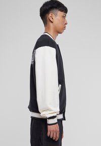 Veste varsity noir et blanc avec des poignets et un col côtelés, présentant un logo sur la poitrine et des poches latérales ; texture de tissu lisse.