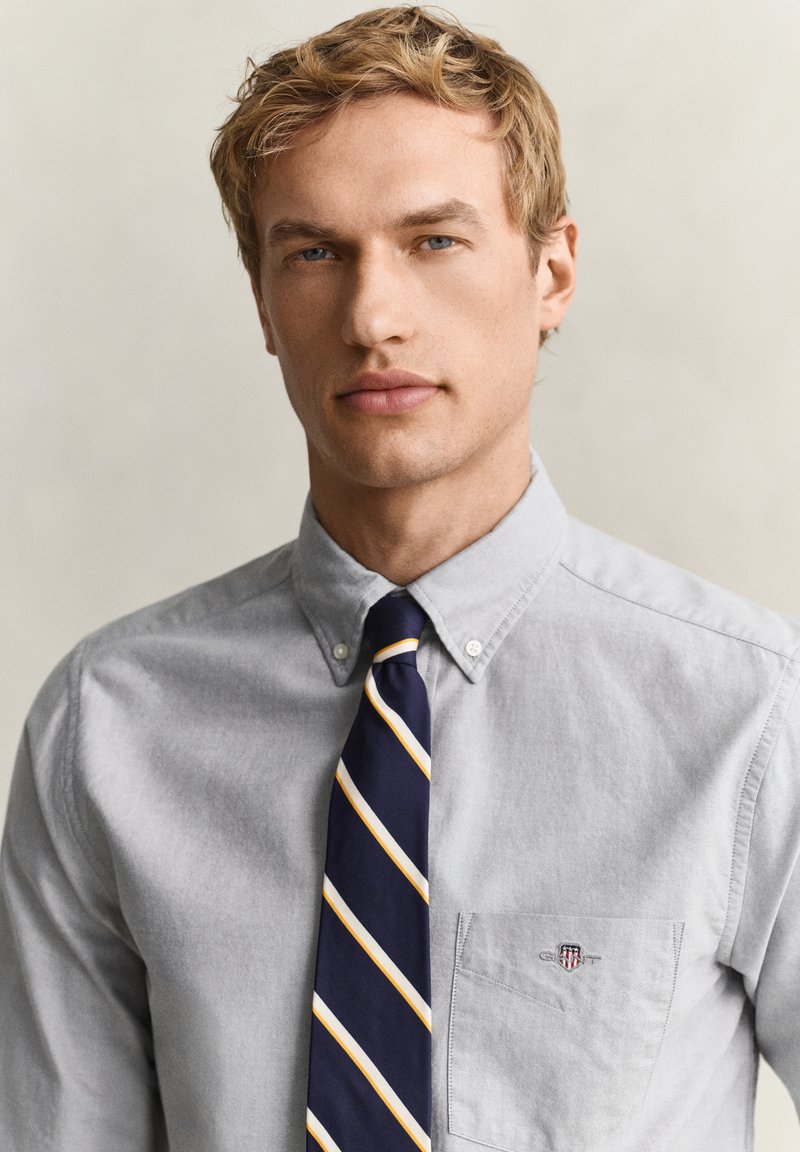 Jeune homme aux cheveux blonds courts portant une chemise gris clair boutonnée et une cravate rayée bleu marine, fond neutre.