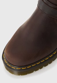 Botte en cuir marron avec un bout arrondi, une surface texturée, des coutures jaunes le long de la semelle et une semelle extérieure en caoutchouc noir robuste.
