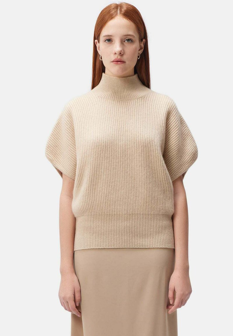 Beige ribbad stickad tröja med hög hals och korta ärmar, med en avslappnad passform och mjuk textur, tillsammans med en beige kjol.