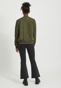 Veste bomber kaki olive avec des poignets et un ourlet côtelés, portée par-dessus un jean évasé noir et des bottes noires. Tissu lisse, design simple, sans motifs.