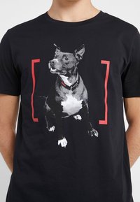 Camiseta negra con un gráfico de un perro en escala de grises, delimitado por corchetes rojos. El perro tiene el pecho blanco y lleva un collar.