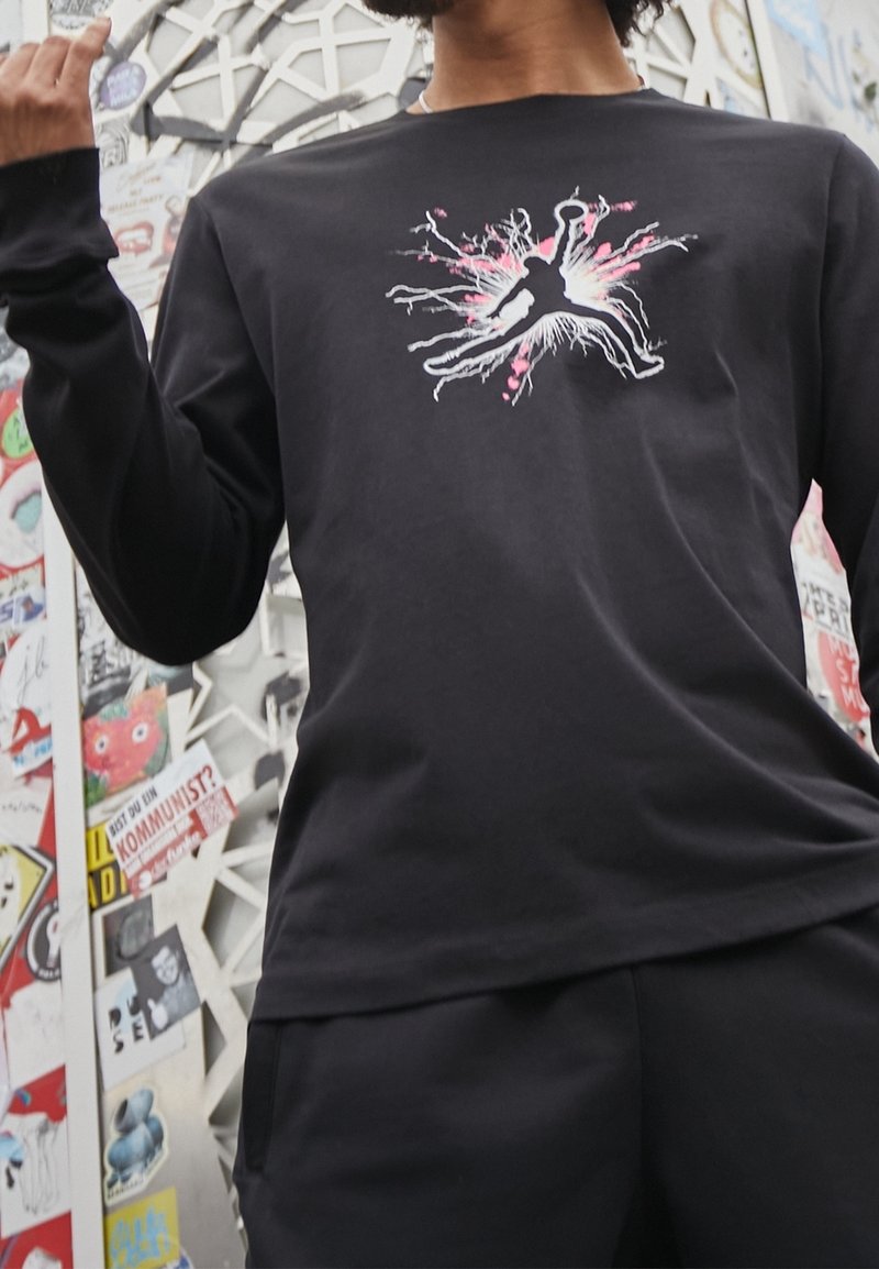 Schwarzes Langarmshirt aus Baumwolle mit einem zentralen Grafikdesign in Pink und Weiß, das eine Figur mit dynamischen Linien zeigt, die nach außen strahlen.