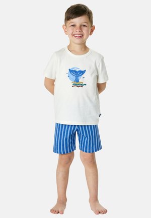 Smiling young boy stands barefoot wearing white t-shirt with blue whale tail graphic and blue striped shorts against white background.