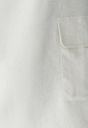 Zbliżenie na beżową kieszeń z widocznymi szwami na teksturowanym, jasnym tle tekstylnym.