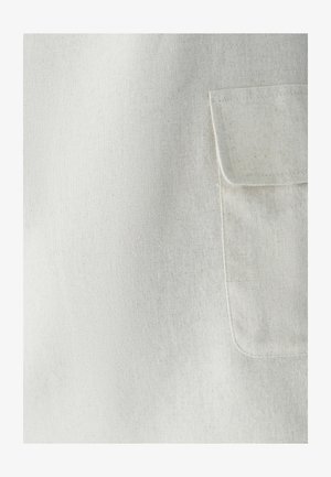 Zbliżenie na beżową kieszeń z widocznymi szwami na teksturowanym, jasnym tle tekstylnym.