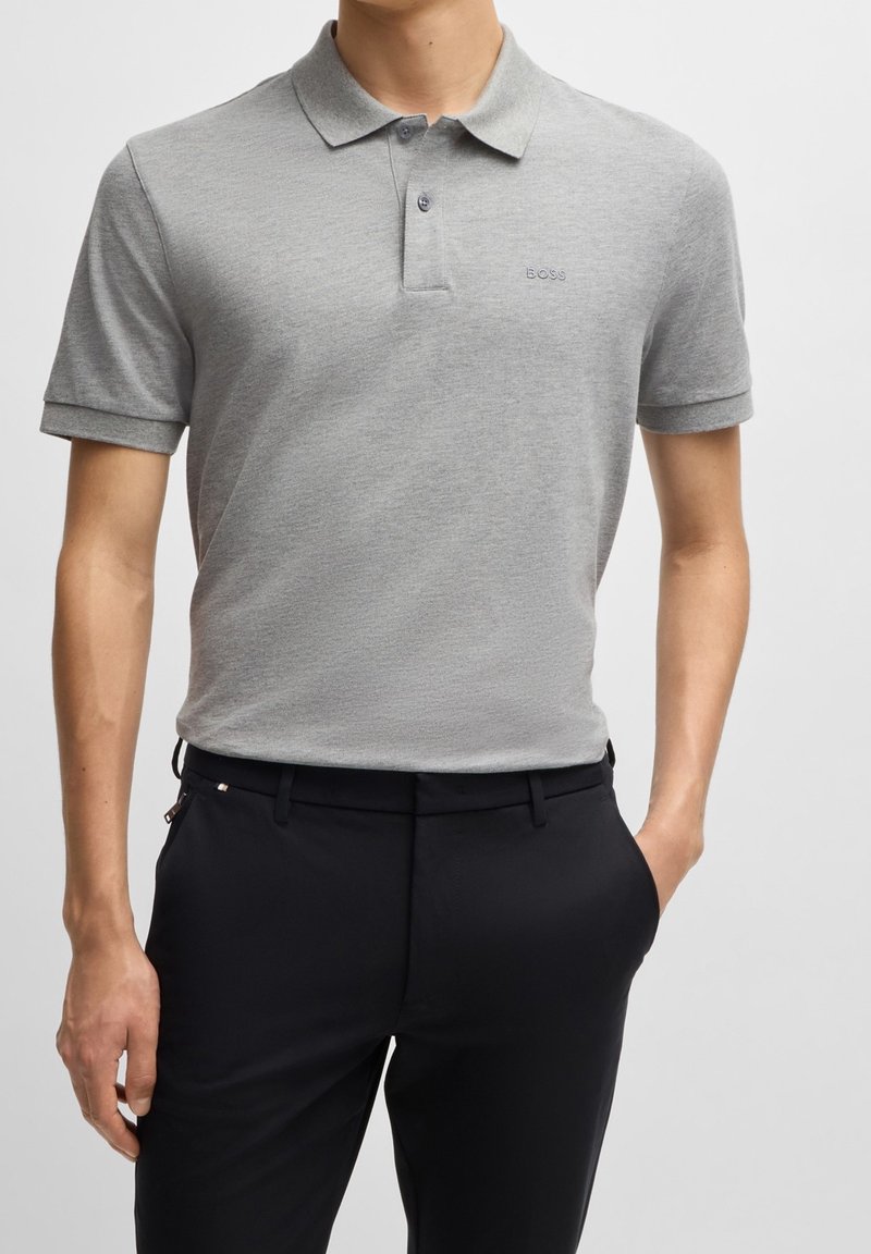 Homme portant un polo gris clair à manches courtes rentré dans un pantalon noir, une main dans la poche, debout devant un fond uni.