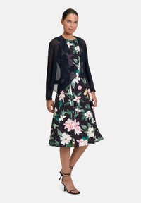Blumenkleid in Midi-Länge mit schwarzem Hintergrund, das große rosa und weiße Blumen zeigt. Kombiniert mit einem transparenten schwarzen Überwurf und schwarzen High Heels.