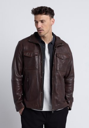 Mann mit kurzen dunklen Haaren, der eine braune Lederjacke über einem weißen Hemd und dunklen Hosen trägt, steht mit den Händen in den Taschen und schaut nach unten.