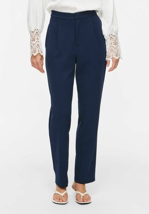 Femme portant un pantalon bleu marine ajusté, un chemisier blanc avec des manches en dentelle et des sandales blanches, se tenant devant un fond uni.