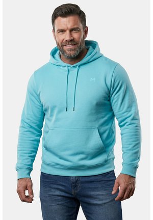 Uomo con la barba che indossa una felpa con cappuccio azzurro chiaro e jeans blu, in piedi contro uno sfondo chiaro e semplice, sorridendo leggermente.