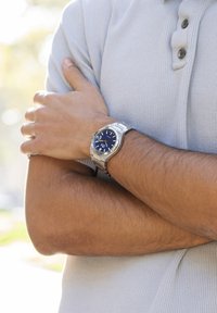 Silverfärgad rostfri stålur med blå urtavla, vita timmarkörer och en polerad bezel, som bärs på ett handled med brun hud, med armarna i kors.