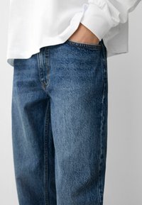 Jeans in denim di un blu medio, con un taglio dritto, lievi sbiaditure e un classico design a cinque tasche, abbinati a una maglietta bianca.