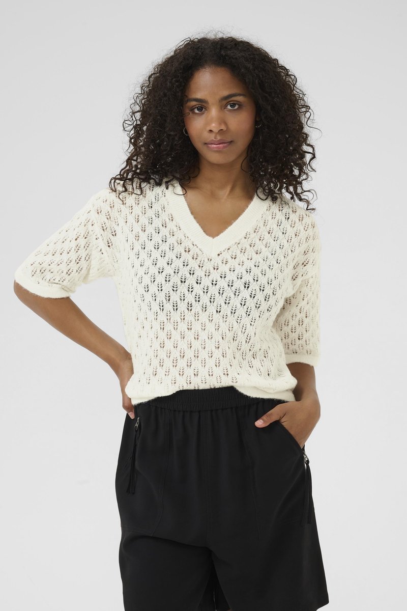 Weißer Strickpullover mit V-Ausschnitt und kurzen Ärmeln, der ein Rautenmuster aufweist. Kombiniert mit schwarzen elastischen Shorts mit Reißverschluss-Taschen.