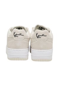 Para kremowych zamszowych sneakersów z grubymi białymi podeszwami, widzianych od tyłu, z czarnym logo w stylu podpisu na każdym obcasie.