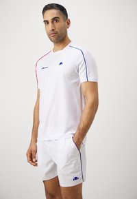 White athletic shirt with red and blue accents, textured fabric, round neckline, paired with white shorts featuring blue piping and logo.