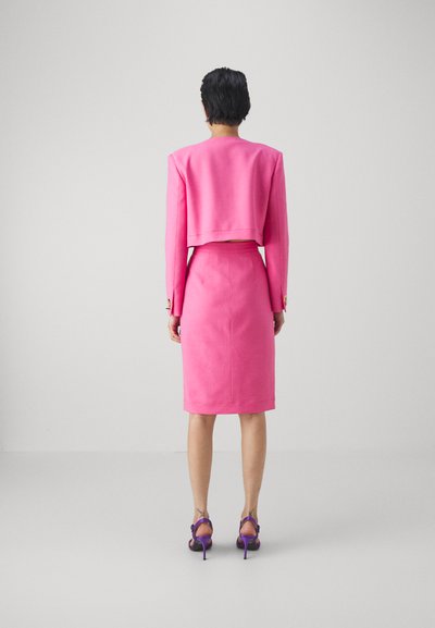 Costume en polyester rose avec une veste courte et une jupe crayon. Présente des accents de boutons dorés et des sandales à talons hauts violettes. Vue de dos.