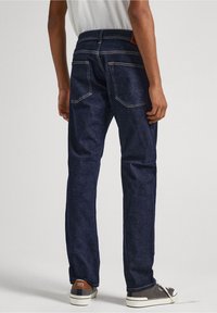 Jeans de mezclilla azul oscuro con corte recto, detalles de costura visibles y dos bolsillos traseros. Llevados con zapatillas grises y una camiseta blanca.