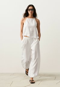 Femme marchant avec un haut blanc ample à col halter et un pantalon blanc large, portant des lunettes de soleil noires et des sandales noires sur un sol en pierre clair.