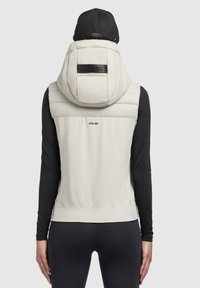 Leichte, cremefarbene Weste mit einer gepolsterten Kapuze, schwarzen Akzenten, geripptem Saum und KHUJO-Logo, getragen über einem schwarzen Langarmshirt.