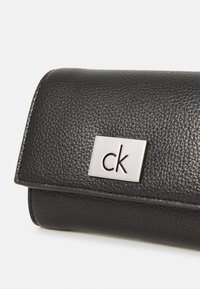 Portafoglio in pelle nera con finitura testurizzata, caratterizzato da una placca con il logo "ck" in argento su un pattino rettangolare. Design compatto ed elegante.