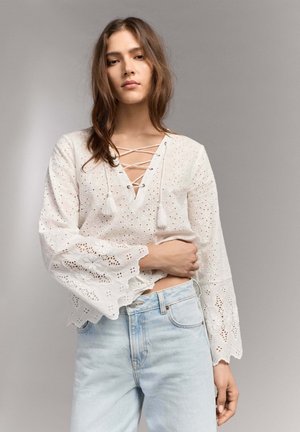 Joven mujer con una blusa blanca de ojales con cordones, mangas florales con borde ondulado, y vaqueros de tiro alto en azul claro, posando frente a un fondo gris.