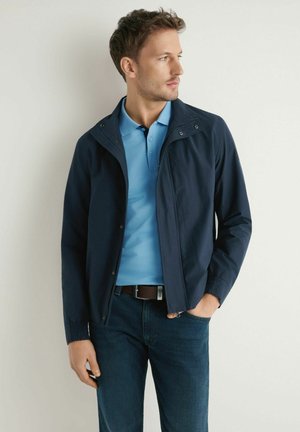 Młody mężczyzna ubrany w granatową kurtkę na jasnoniebieskiej koszulce polo i ciemnych dżinsach, stojący na tle jednolitej białej ściany, patrzący w bok.