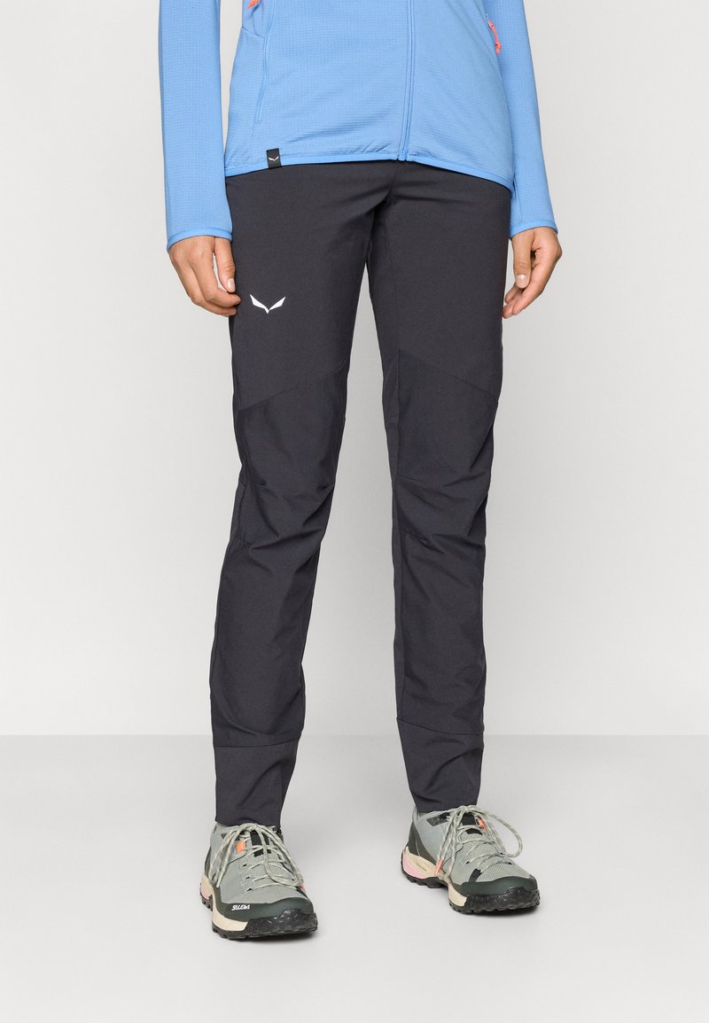 Μαύρα παντελόνια εξωτερικού χώρου με αντίθετα πάνελ, κατασκευασμένα από ανθεκτικό ύφασμα. Συνδυάζονται με γκρι μποτάκια πεζοπορίας με πορτοκαλί λεπτομέρειες.