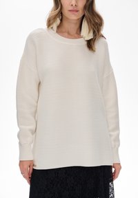 Kobieta w dużym, kremowym swetrze z prążkowanym splotem i długimi rękawami, w czarnej spódnicy z koronkowymi kwiatami, w zestawieniu z dużymi, kremowymi chwostami w uszach.