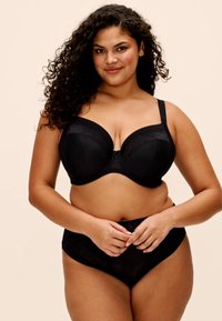 Femme aux cheveux bouclés portant un soutien-gorge noir et une culotte taille haute, debout et souriante devant un fond beige uni.