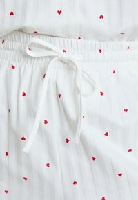 White fabric with vertical stripes, featuring scattered red heart patterns. Elastic waistband with a tied drawstring detail.