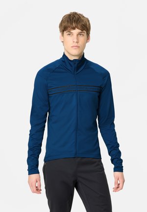 Jeune homme portant une veste bleue zippée avec des bandes noires sur la poitrine et un pantalon noir, debout devant un fond uni.