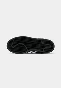 Semelle de chaussure de sport noire avec un motif de traction en zigzag, comprenant des accents blancs et une surface texturée pour une meilleure adhérence.