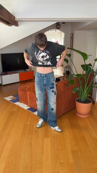 Niebieskie dżinsy z efektem przetarć i wzorami z naszywkami, o luźnym kroju, z licznymi rozdarciami i zapinane na guziki. Osoba nosi czarny t-shirt z grafiką czaszki.