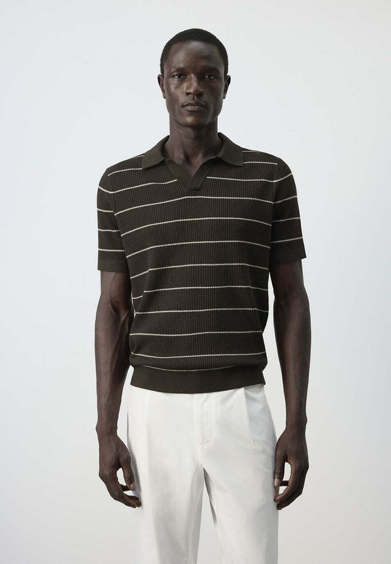 Homme portant un pull en tricot à manches courtes vert foncé avec des rayures blanches et un pantalon blanc, debout devant un fond clair uni.