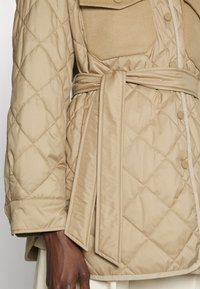 Veste matelassée beige avec taille ceinturée, dotée d'une poche poitrine en daim, de boutons-pression et d'un tissu texturé.