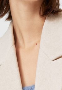 Abrigo de lana beige con cuello con muesca, textura suave y un lunar oscuro visible en la zona del cuello, llevado sobre una prenda azul claro.
