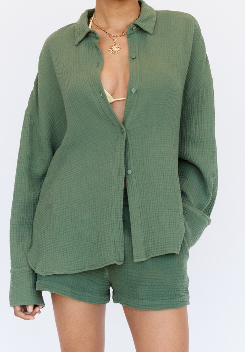 Femme portant une chemise boutonnée ample texturée vert olive et un short assorti, avec un collier en forme de soleil et un haut de bikini visible en dessous.