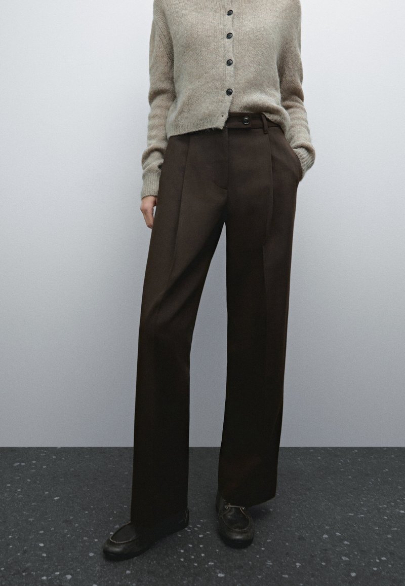 Personne portant un cardigan beige boutonné, un pantalon large marron foncé et des chaussures noires, debout contre un mur uni gris clair.