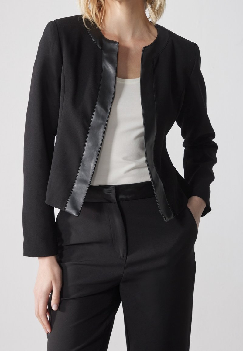 Black blazer with a round neck, contrasting leather trim, structured shoulders, and a tailored fit, paired with black trousers.