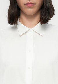 Camisa de colarinho branco feita de tecido suave; possui uma frente abotoada com pequenos botões brancos redondos e um colarinho clássico e estruturado.