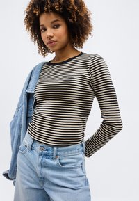 Giovane donna con capelli ricci che indossa una maglietta a maniche lunghe a righe nere e beige, jeans azzurri e tiene una giacca di denim su una spalla.